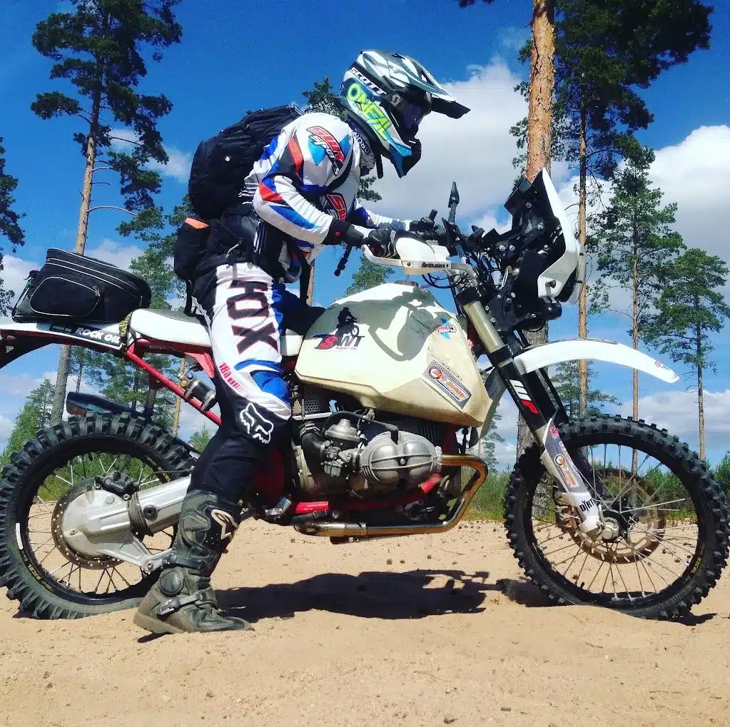 Motorradfahrer auf umgebauter BMW R 100 R mit großem Wüstentank im Sand, umgeben von Kiefern und blauem Himmel.