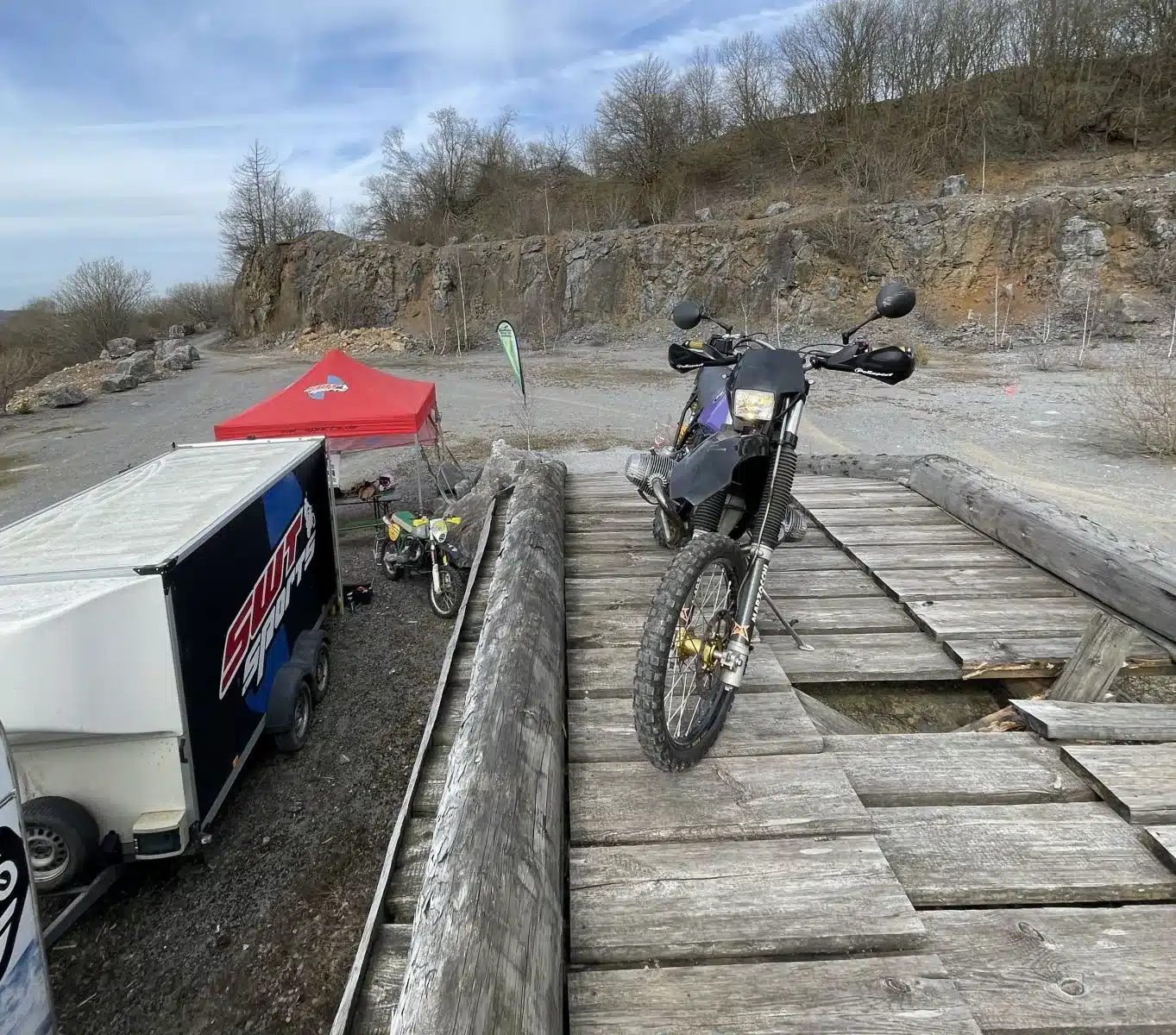 SWT-SPORTS EnduroBoxer Nr. 150, eine modifizierte BMW R 80 G/S, abgestellt auf einem Holzbalkensteg vor einem Offroad-Gelände mit Anhänger und Zelt im Hintergrund.