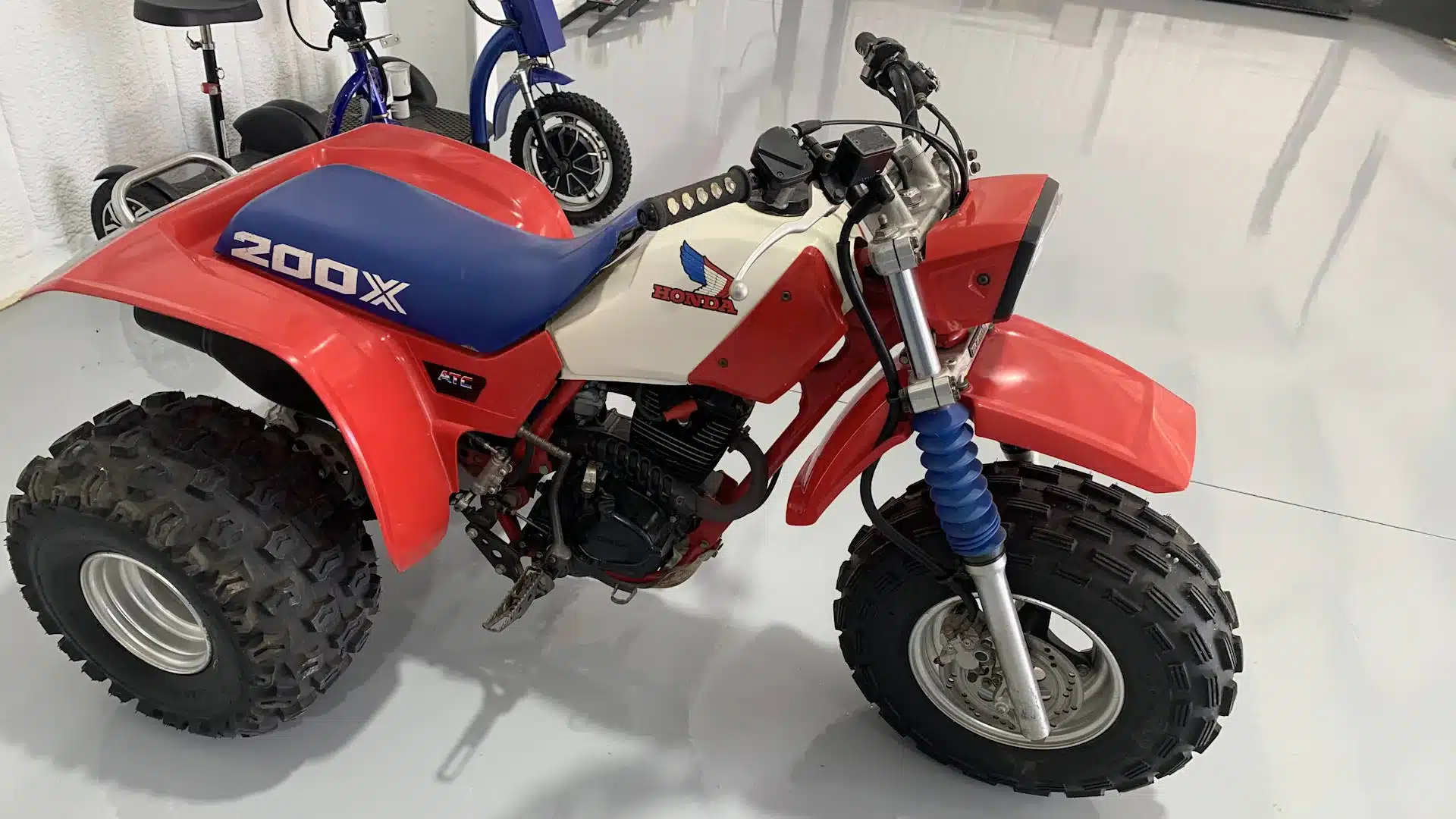 Honda ATC 200X von 1985 mit roten Plastikteilen, weißem Tank und blauer Sitzbank in einer Halle, seitlich aufgenommen.