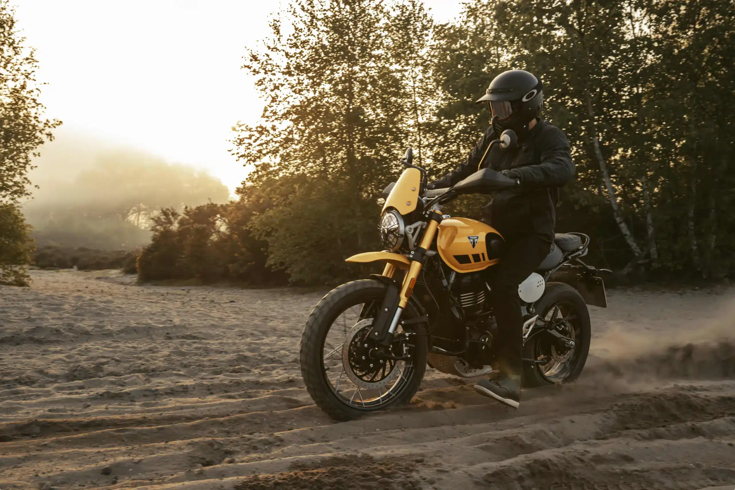 Triumph Scrambler 400 XC in gelb fährt auf sandigem Untergrund durch eine bewaldete Landschaft