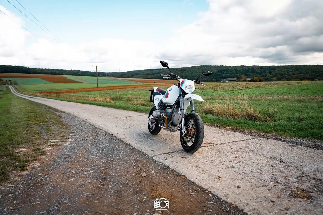 Eine SWT-SPORTS Supermoto BMW R 1043 SM steht auf einer schmalen Landstraße mit Feldern und Wald im Hintergrund